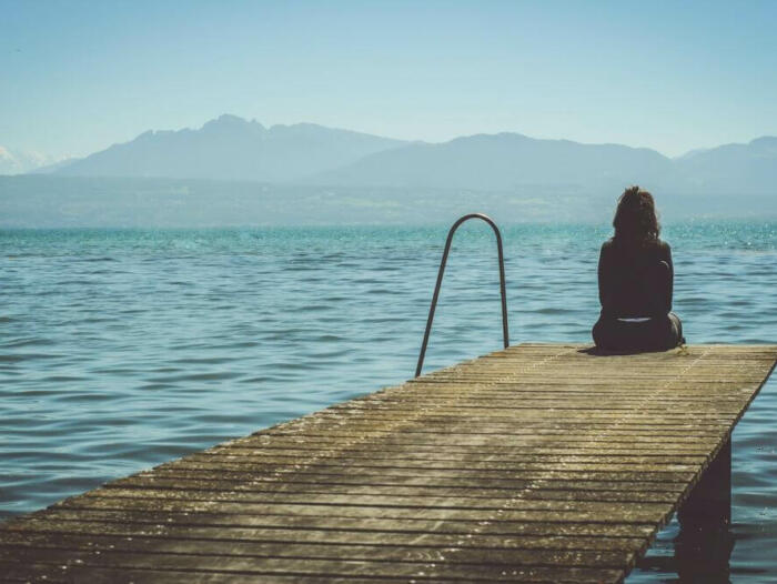 a woman sits on the end of a dock during daytime staring across a lake